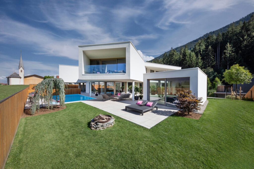 Moderne weiße Villa mit Pool, Terrasse und großzügigem Garten vor Bergkulisse.