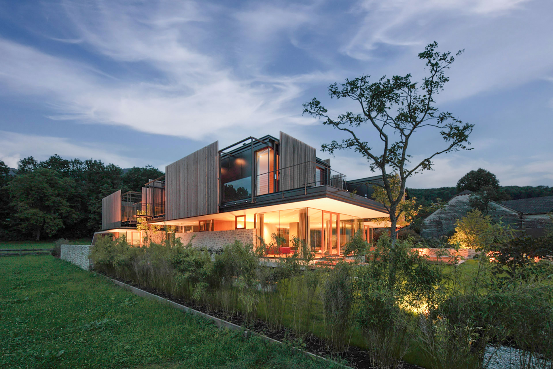 Moderne Villa bei Dämmerung, mit Holzlamellen-Fassade, großen Glasflächen und Garten im Vordergrund.