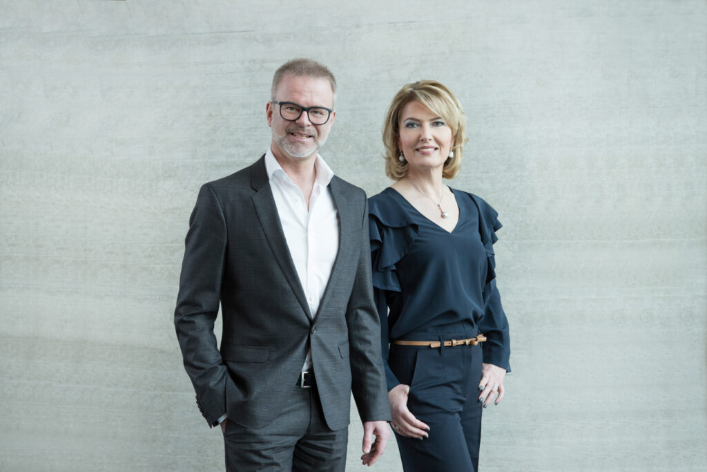 Mann und Frau im Business-Outfit vor Betonwand.