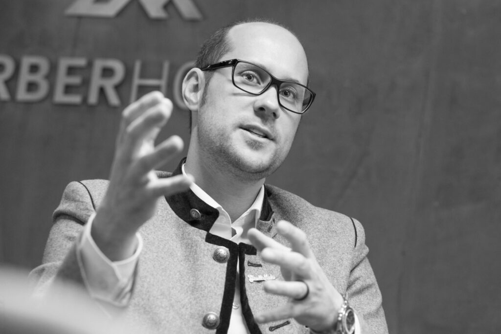 Nahaufnahme eines entspannten Mannes mit hellem Hemd und freundlichem Ausdruck, aufgenommen in einem modernen Innenraum.