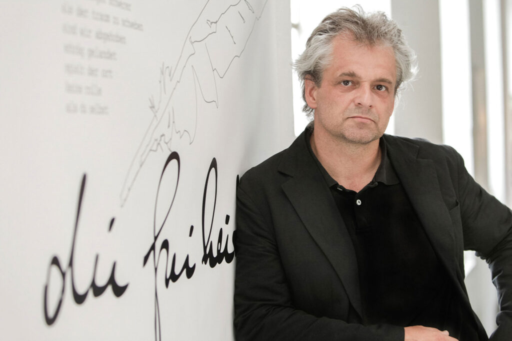 Mann mit grauen Haaren im schwarzen Sakko vor einer beschrifteten Wand.