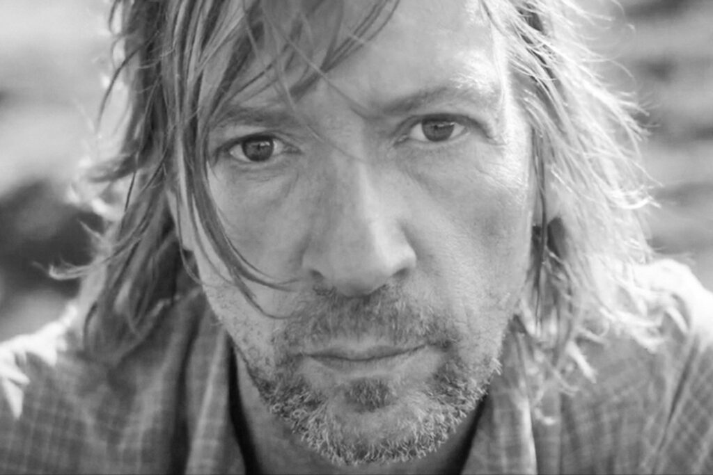 Porträt eines Mannes mit mittellangen Haaren und Bart, der direkt in die Kamera blickt.