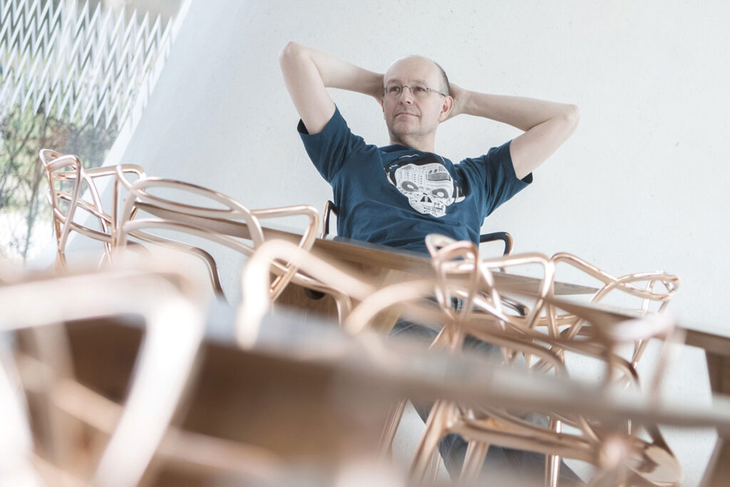 Mann lehnt entspannt auf Stuhl zwischen gestapelten Holzstühlen.