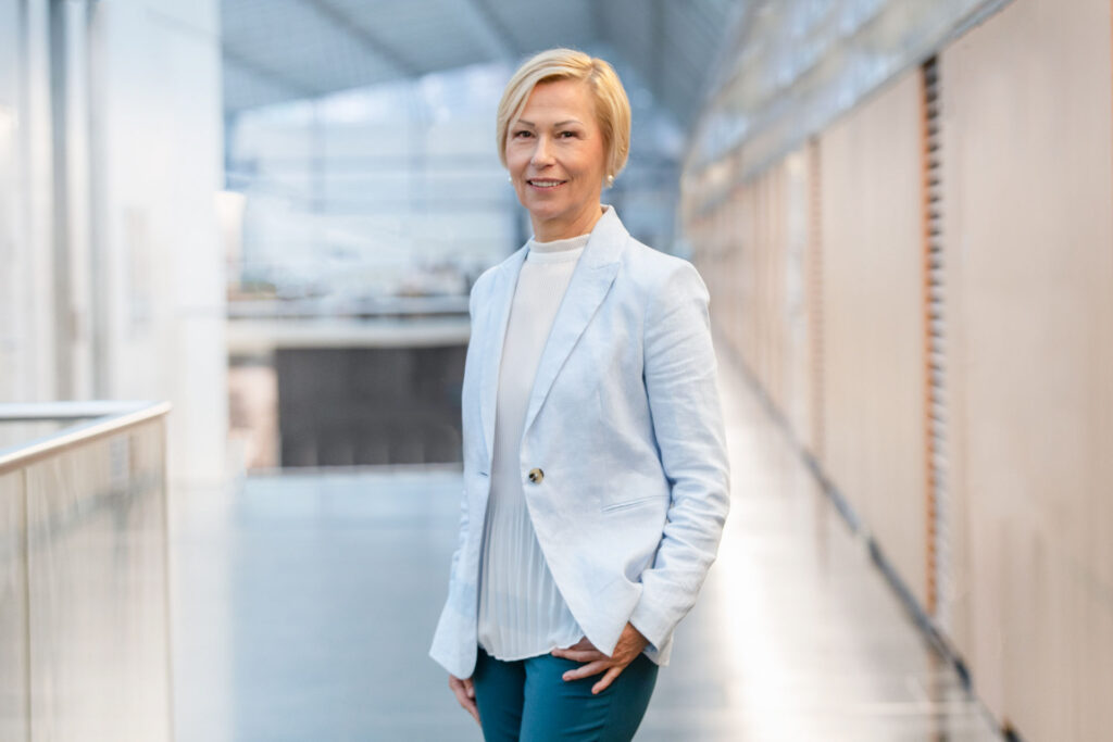 Frau im hellblauen Blazer steht in modernem, lichtdurchflutetem Gebäude.