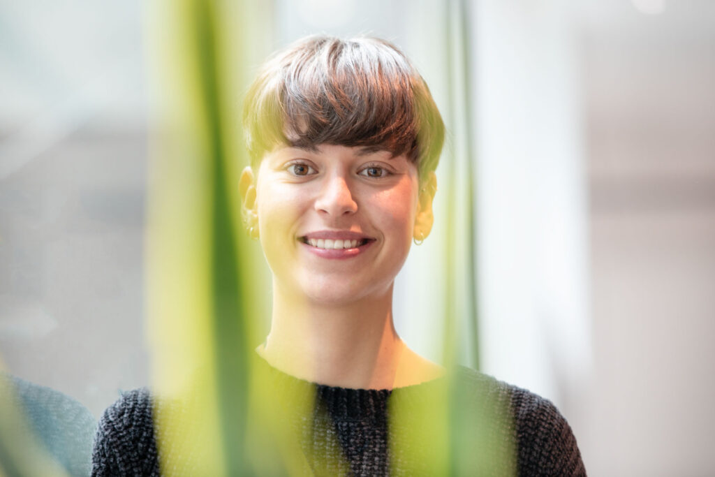 Junge Frau mit kurzem Haarschnitt lächelt in die Kamera, grüne Pflanzen im Vordergrund.