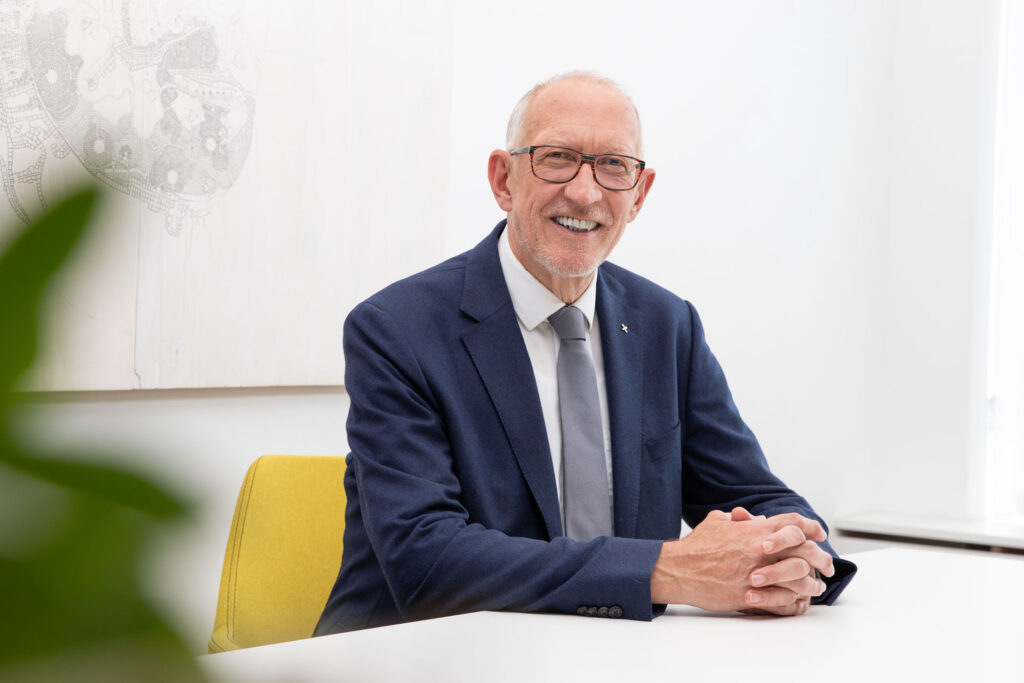 Älterer Mann im Anzug sitzt lächelnd an einem Tisch.