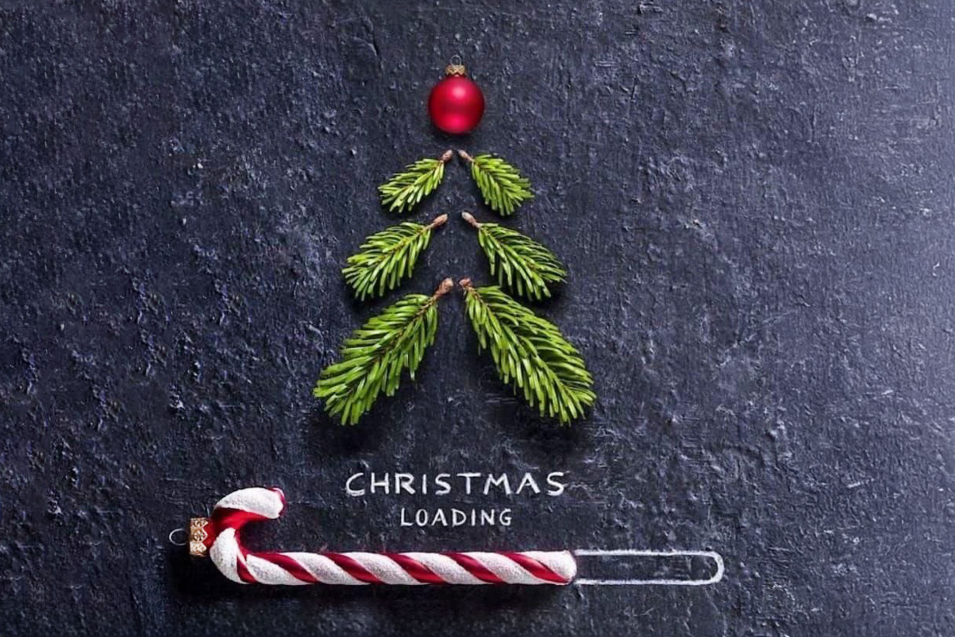 Minimalistisches Weihnachtsmotiv aus Tannenzweigen, roter Kugel und Zuckerstange auf dunklem Hintergrund mit dem Schriftzug ‚Christmas Loading